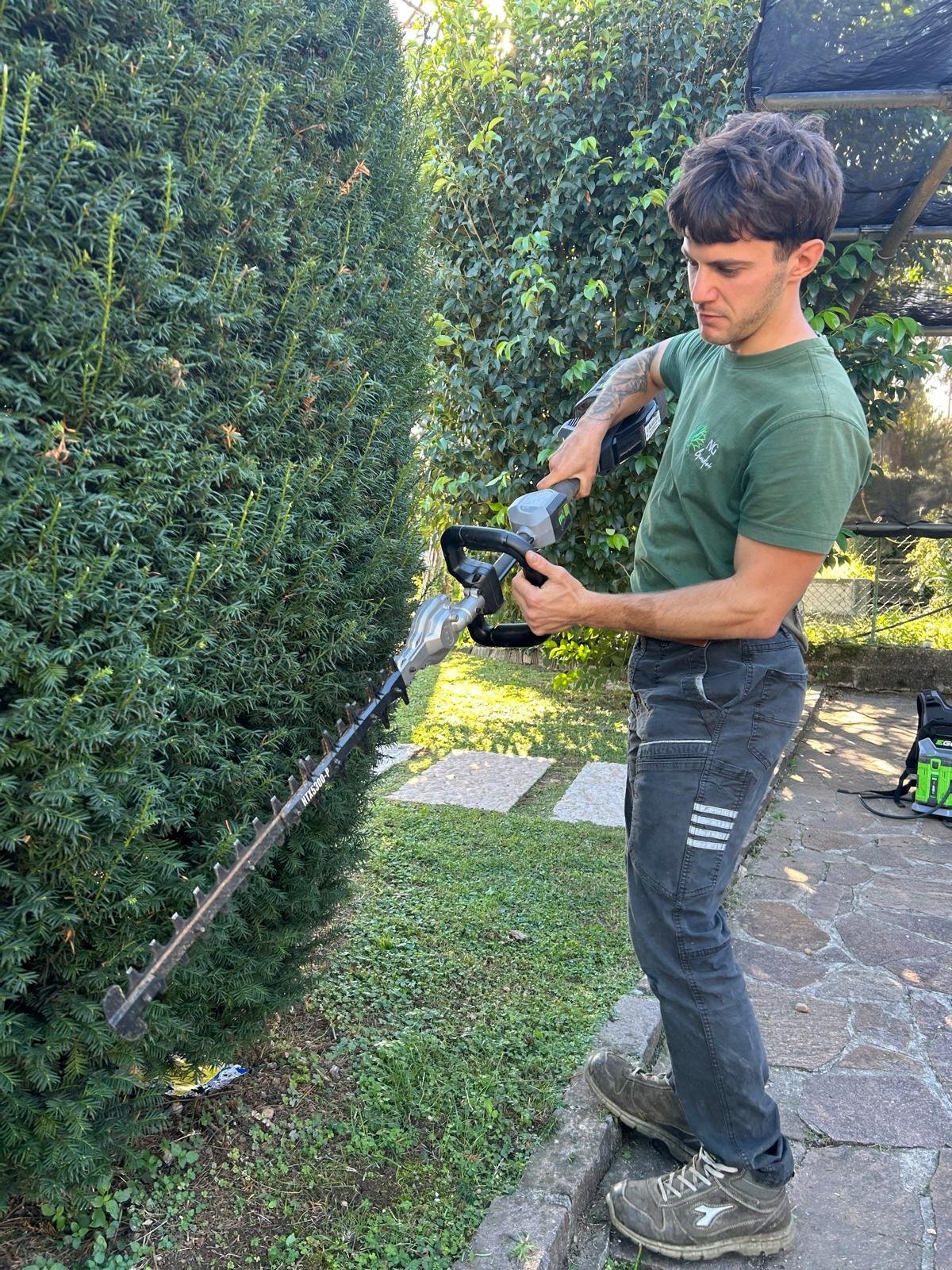 Operatore NG Giardini in maglietta verde che pota una siepe alta con tagliasiepi a batteria
