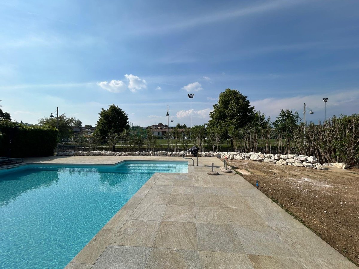 Piscina esterna con acqua blu, deck in cemento e alberi sotto il cielo azzurro
