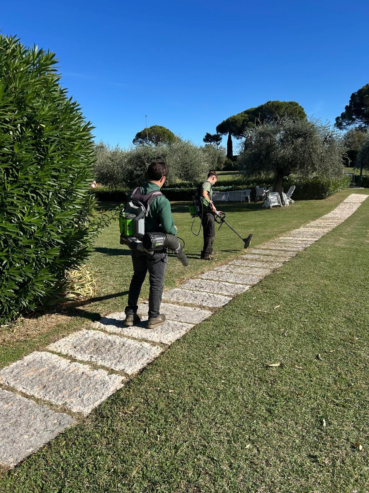 Due operatori con soffiatore e decespugliatore a batteria sul prato curato accanto a un vialetto in pietra
